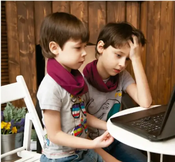 Dos niños pequeños sentados frente a una computadora portátil sobre una mesa redonda, aparentemente compartiendo una actividad digital en un ambiente con paredes de madera.