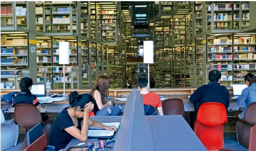 Fotografía de una biblioteca con estantes llenos de libros al fondo y varias personas sentadas en mesas de estudio trabajando con computadoras y cuadernos, iluminadas por lámparas blancas.