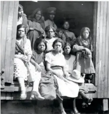 Fotografía en blanco y negro de un grupo de mujeres y niñas sentadas en la puerta de un vagón de tren, con bultos y enaguas, mirando a la cámara.
