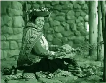 Fotografía a color (tono verdoso por la impresión) de una mujer adulta sentada en el suelo frente a un muro de piedra, tejiendo en un telar; viste poncho rojo con patrones intrincados y sombrero o montera con bordados multicolores. Alrededor hay tejidos en el piso.