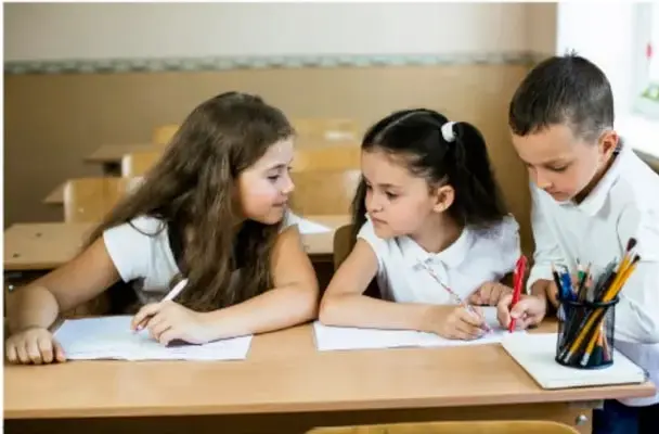 Tres estudiantes de primaria trabajando en equipo sobre una mesa con hojas y lápices.