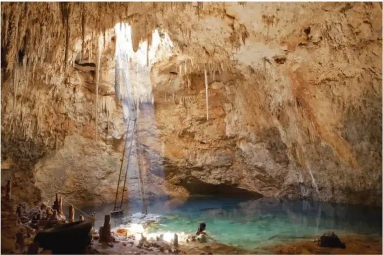 Fotografía de un cenote en Cuzamá, Yucatán: caverna con estalactitas y estalactitas colgantes, un haz de luz solar que entra por una abertura superior e ilumina el agua turquesa; a la orilla se observan rocas, una pequeña embarcación y una estructura de madera.