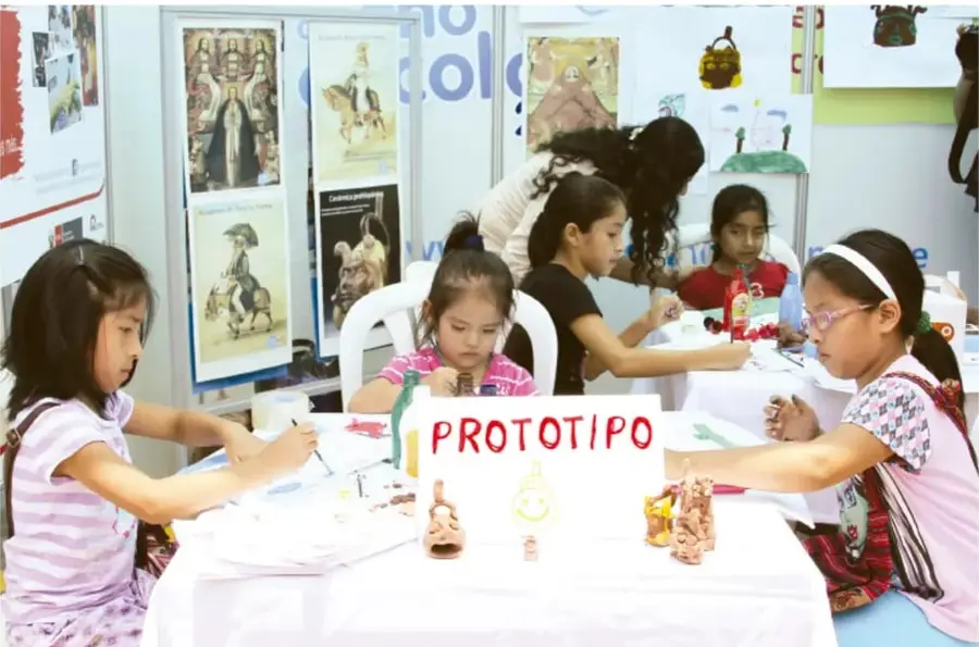 Fotografía de niñas de primaria trabajando en una mesa con un letrero que dice PROTOTIPO y varias figuras de barro o plastilina; al fondo hay carteles escolares.