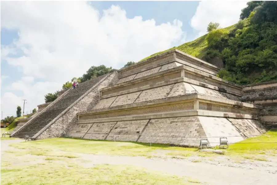 Fotografía de la Gran Pirámide de Cholula (Tlachihualtépetl), vista desde un costado. Se observa una construcción escalonada de piedra con una gran escalinata central, rodeada de vegetación y césped, bajo un cielo con nubes.