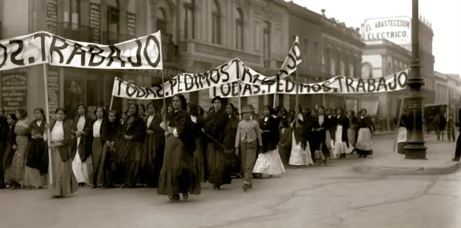 Fotografía histórica en sepia de una manifestación de mujeres con mantones sobre los hombros portando pancartas que dicen 'PEDIMOS TRABAJO' y 'TODAS PEDIMOS TRABAJO', marchando por una avenida empedrada de una ciudad mexicana con edificios del porfiriato al fondo; pie de foto: Manifestación de mujeres en demanda de trabajo, 1913.