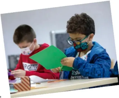 Fotografía de dos niños en un salón de clases; uno de ellos sostiene una hoja verde y el otro trabaja con materiales sobre la mesa.