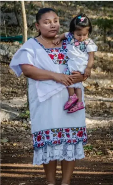 Fotografía de una mujer maya con vestido blanco tradicional bordado, cargando a una niña pequeña a horcajadas sobre su cintura durante la ceremonia del jestsméek'.