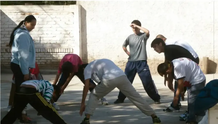 Grupo de estudiantes de secundaria realizando estiramientos y ejercicios de calentamiento al aire libre en una cancha con muro de ladrillo; un integrante observa protegiéndose los ojos del sol.