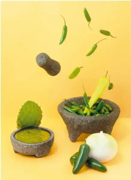 Fotografía sobre fondo amarillo de un molcajete de piedra con chiles verdes, un tejolote volando, chiles en el aire, un molcajete pequeño con salsa verde, una penca de nopal, una cebolla blanca y chiles serranos.