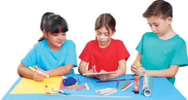 Tres estudiantes (dos niñas y un niño) sentados a una mesa azul construyen una maqueta con hojas de papel, plasticina, palos de helado, tubos de cartón y témperas, entre risas y colaboración.