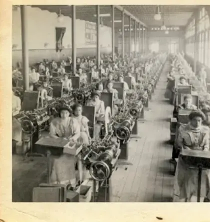 Fotografía antigua en blanco y negro de mujeres trabajando en una fábrica textil, sentadas frente a máquinas de coser.
