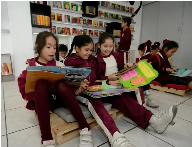 Estudiantes de primaria con uniforme escolar leen libros sentados en tarimas de madera dentro de una biblioteca.