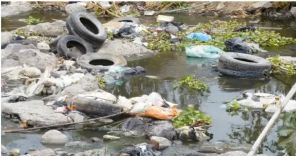 Fotografía de un cuerpo de agua contaminado con basura, llantas y desechos plásticos flotando, rodeado de vegetación.