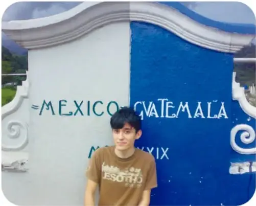 Adolescente de playera café sonriendo frente a un letrero pintado de blanco y azul que lee 'MÉXICO GUATEMALA' con una fecha inferior no del todo legible.