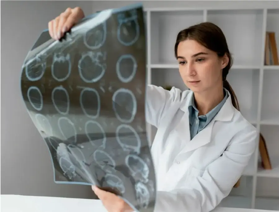 Profesional de la salud (médica o radióloga) con bata blanca observando una placa radiográfica con múltiples imágenes de cortes de cerebro (estudio de tomografía/resonancia).