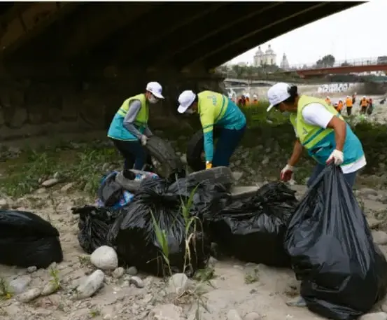 Fotografía de voluntarios con chalecos y gorras recogiendo residuos en bolsas negras en una zona a la orilla de un río o bajo un puente, acción comunitaria de limpieza.