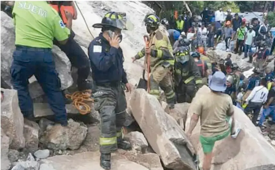 Fotografía de un grupo de rescatistas y voluntarios, algunos con uniforme y casco, trabajando entre grandes rocas y escombros; al fondo se observan más personas y una zona de deslave.