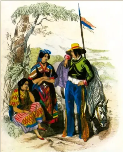 Grabado coloreado titulado «Mestizos y guerrilleros de Michoacán», publicado en Viajes alrededor del mundo y naufragios célebres, París, 1814. Muestra a un grupo de personas indígenas y mestizas junto a un árbol: mujeres sentadas con rebozos y faldas tradicionales coloridas y un hombre de pie con sombrero de ala ancha, saco verde, pantalones azules, sable al cinto y un estandarte o bandera, junto a un animal (burro o caballo). Acervo INEHRM.