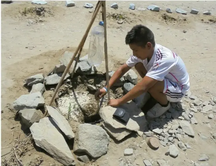 Fotografía de un estudiante trabajando en una construcción de piedras al aire libre, sosteniendo una herramienta, con una estructura de palos y una botella de plástico colgada; representa el trabajo práctico y comunitario del proyecto.
