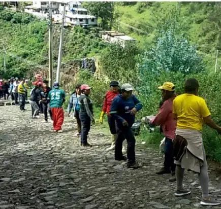 Fotografía de un grupo de personas caminando por un camino empedrado en una zona rural montañosa,许多人 con chalecos de trabajo, posiblemente realizando una jornada de trabajo comunitario o tequio.