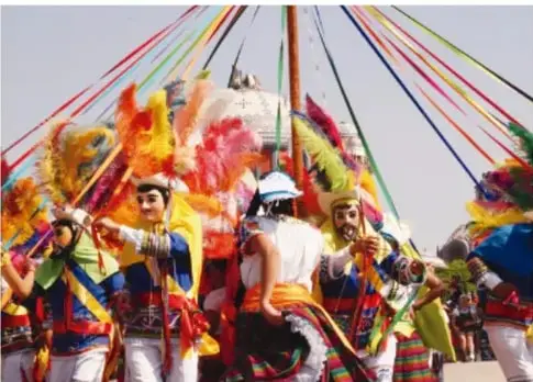 Danza tradicional con danzantes cubiertos con capas de colores y máscaras, uno de pie y otro arrodillado, en una plaza empedrada.