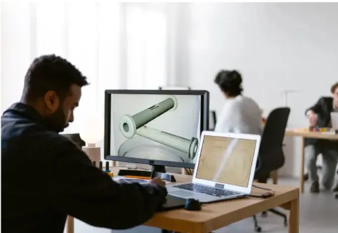 Fotografía de un oficina donde un hombre trabaja frente a una computadora con software de diseño CAD de una pieza mecánica; otras personas trabajan al fondo con laptops.