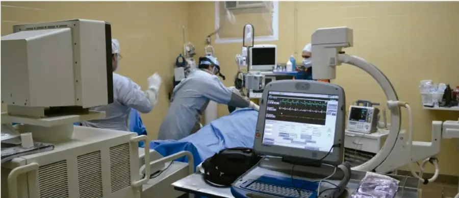 Imagen de un quirófano o sala de intervención con personal médico en batas y mascarillas, monitores que muestran signos vitales, equipo de rayos X tipo C y computadoras, ilustrando la tecnología aplicada a la atención médica.