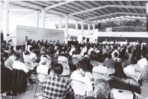 Fotografía en blanco y negro de una reunión masiva de docentes en un recinto techado con techo metálico y estructura de vigas; al fondo, una presidencia con manta y pantalla. El público numeroso está sentado en sillas plegadizas de espaldas a la cámara.