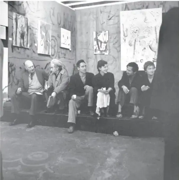 Fotografía en blanco y negro de un grupo de jóvenes escritores sentados en una banca dentro de un espacio con dibujos y carteles en las paredes.