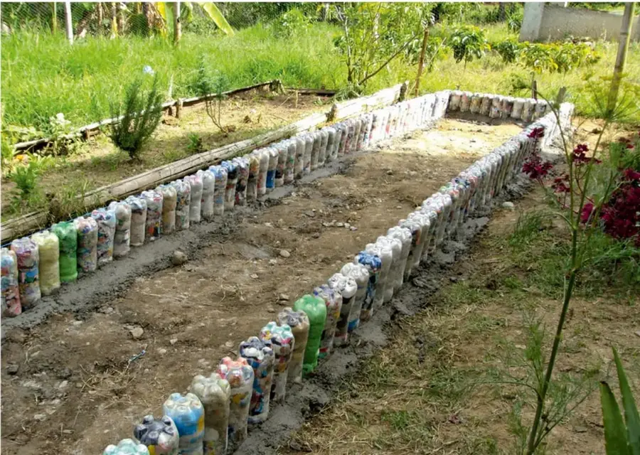 Muro rectangular construido con botellas de plástico rellenas y compactadas, unidas con mezcla de cemento, que delimita un espacio de tierra para cultivo; alrededor hay pasto, plantas y flores.