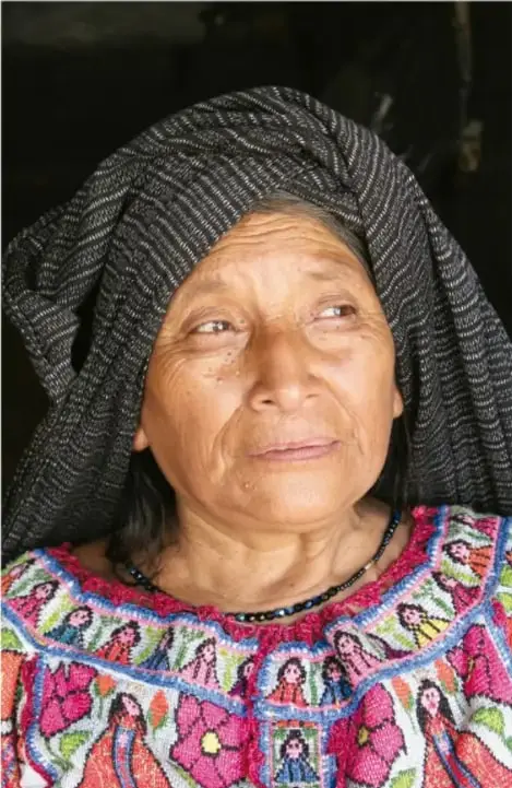 Retrato de una mujer indígena adulta con rebozo gris estampado y huipil bordado de colores vivos, posiblemente artesana o participante de una comunidad.