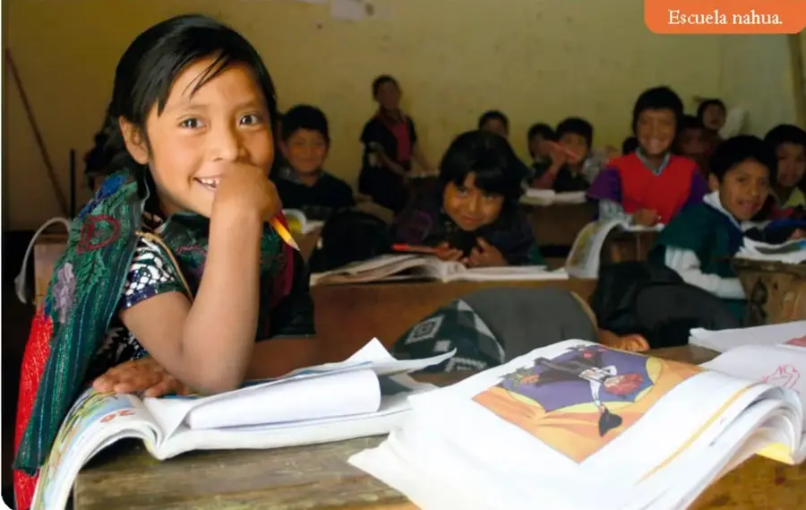 Niña indígena sonriente en un salón de clases con varios compañeros y libros abiertos; etiqueta 'Escuela nahua'.
