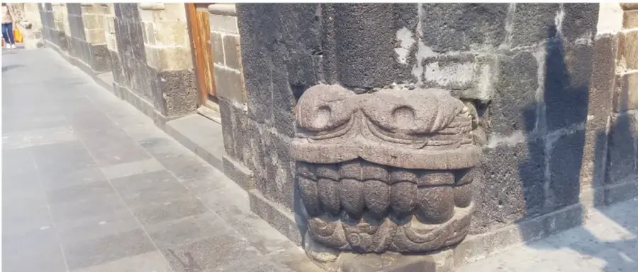 Fotografía cercana de la cabeza de serpiente de piedra en la esquina del edificio: se distinguen sus grandes dientes, ojos y fauces tallados en piedra volcánica, junto a una acera de losas.