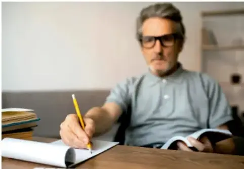 Fotografía de un hombre adulto con lentes escribiendo con lápiz en un cuaderno sobre una mesa, junto a otros libros.