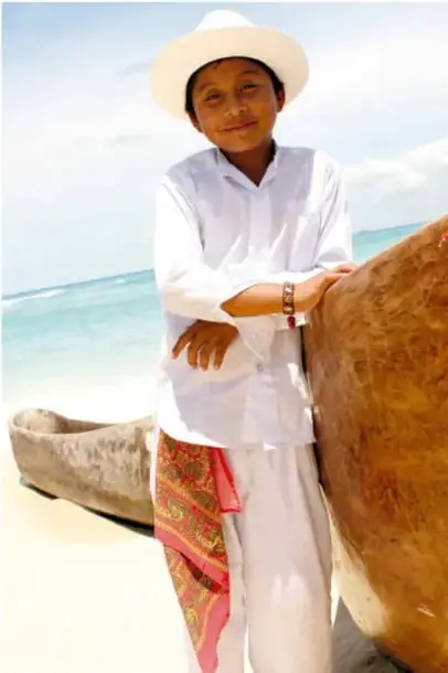 Fotografía de una persona con sombrero de ala ancha y ropa blanca junto a una canoa en la playa, ilustrando la diversidad cultural y lingüística.