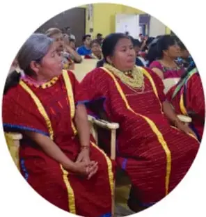 Fotografía circular de mujeres indígenas de traje tradicional rojo con collares, sentadas en una asamblea o reunión comunitaria.