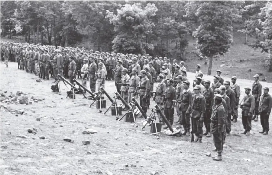 Fotografía en blanco y negro de una formación militar al aire libre: numerosos soldados uniformados y con cascos, alineados en filas sobre un terreno pedregoso, con morteros y equipo militar en primer plano; al fondo se observan árboles y colinas.