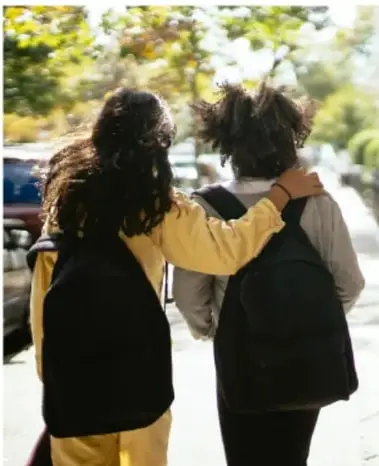 Fotografía de dos estudiantes con mochilas caminando juntos por la calle, uno con el brazo sobre el hombro del otro, en un entorno arbolado y soleado.