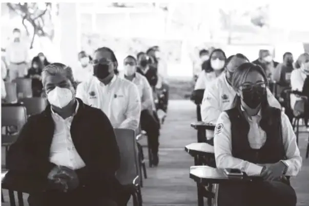 Fotografía en blanco y azulado de adultos (aparente formación docente) sentados en sillas con bloque de escritura, varios con cubrebocas.