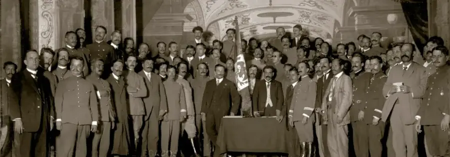 Fotografía histórica en sepia de un gran grupo de hombres —delegados militares y civiles— posando formalmente alrededor de una mesa con un pizarrón, en la Soberana Convención de Aguascalientes, 10 de octubre de 1914.