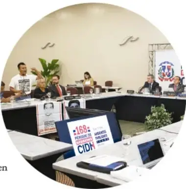Fotografía circular de una asamblea o mesa de trabajo con participantes sentados frente a laptops; hay carteles con lemas como '168 años de abolición de la esclavitud' y logotipos de organismos de derechos humanos.
