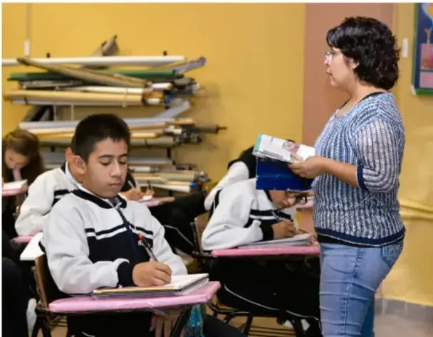 Fotografía de un aula: una docente de pie entrega libros mientras alumnos de secundaria con uniforme escriben en sus cuadernos.