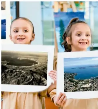 Dos niñas sonrientes sostienen fotografías grandes de paisajes (una aérea de un cráter y otra de una costa) para la exposición.