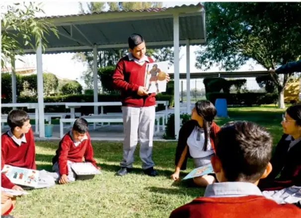 Fotografía de un grupo de estudiantes de telesecundaria con uniforme rojo reunidos en el patio de su escuela; una estudiante de pie lee o presenta un documento mientras los demás, sentados en el pasto, escuchan con sus materiales.