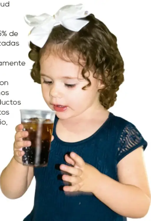 Fotografía de una niña pequeña con moño blanco bebiendo un refresco de cola con popote.