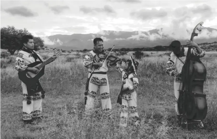 Fotografía en blanco y negro de cuatro personas con trajes tradicionales bordados tocando instrumentos musicales (guitarra, violín, guitarra pequeña y contrabajo) en un campo abierto con montañas al fondo.