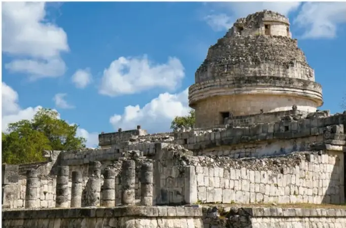Fotografía de una construcción prehispánica de piedra con una torre semicircular en la parte superior, identificada como el observatorio maya de Chichén Itzá (El Caracol), bajo un cielo azul con nubes.