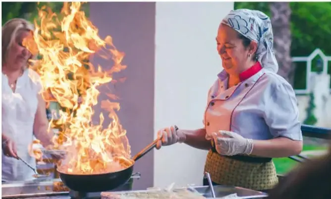 Fotografía de dos personas cocinando con un sartén sobre una llama alta; se observa el fuego y el mango del utensilio.