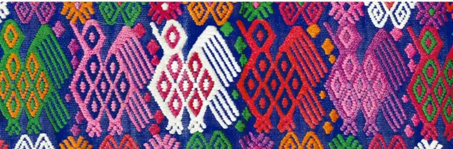 Fotografía de un tejido tradicional mexicano muy colorido, con figuras geométricas y aves bordadas en azul, rosa, verde, rojo y naranja.