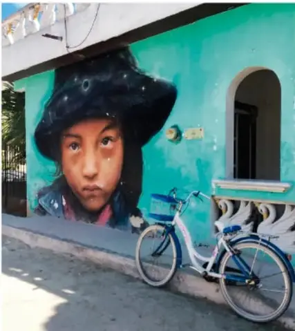 Mural urbano de una niña con sombrero negro sobre pared de color turquesa, con una bicicleta azul apoyada al frente; fotografía de calle con arte comunitario.